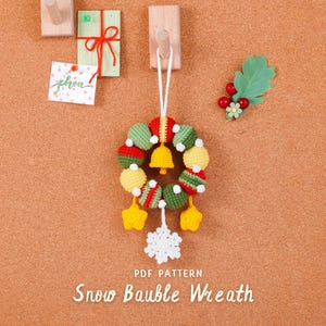 Könnte beinhalten: Ein handgefertigter gehäkelter Weihnachtskranz mit roten, grünen und gelben Kugeln, einer goldenen Glocke und weißen Schneeflocken- und Sternakzenten. Der Kranz hängt an einer weißen Schnur. Der Text "Snow Bauble Wreath" steht unten.