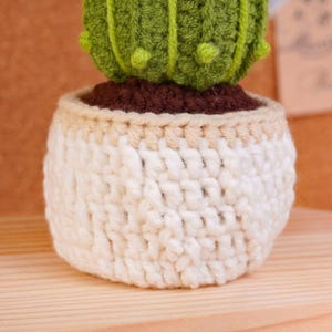 May include: A crocheted cactus in a small pot. The cactus is bright green with vertical ridges and small raised bumps. It sits in a dark brown soil-like base, which is nestled inside a textured, off-white crocheted pot with a light tan rim.