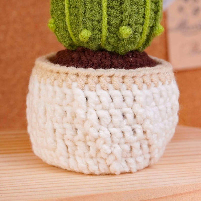 May include: A crocheted cactus in a small pot. The cactus is bright green with vertical ridges and small raised bumps. It sits in a dark brown soil-like base, which is nestled inside a textured, off-white crocheted pot with a light tan rim.