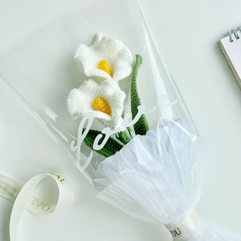 May include: A bouquet of white crocheted calla lilies with yellow centers, wrapped in white tulle and clear plastic with the word "love" written on the plastic.