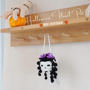 Könnte beinhalten: Ein Holzregal mit Haken, beschriftet mit "Halloween Floral Pot PDF Pattern". An einem der Haken hängt ein gehäkelter Schädel mit lila Blumen und schwarzen Haaren.