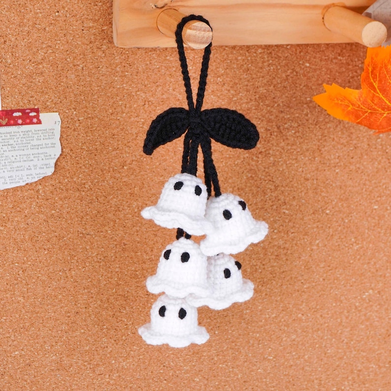 Peut inclure: D&eacute;coration d'Halloween faite &agrave; la main avec cinq petits fant&ocirc;mes blancs crochet&eacute;s avec des yeux &agrave; points noirs. Les fant&ocirc;mes sont suspendus &agrave; un n&oelig;ud et une boucle noirs crochet&eacute;s. Le fond est un tableau en li&egrave;ge.
