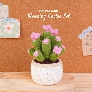May include: A crocheted cactus in a white and beige pot with pink flowers, featuring the text PDF PATTERN Blooming Cactus Pot. The cactus is green with dark brown soil visible at the base, and the pot has a textured, stitched appearance.