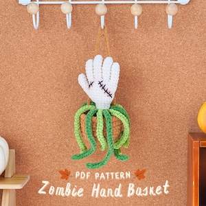 May include: A crocheted zombie hand basket with green and brown yarn. The hand is white with stitched-on stitches. The basket is hanging from a hook on a corkboard wall. The text "PDF PATTERN Zombie Hand Basket" is below the basket.