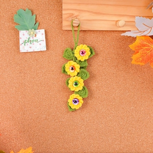 May include: A handmade crochet floral ornament featuring four yellow flowers with colorful centers and green leaves. The ornament is suspended by a green string and is attached to a wooden surface. A small card with the word "phon" is also visible.