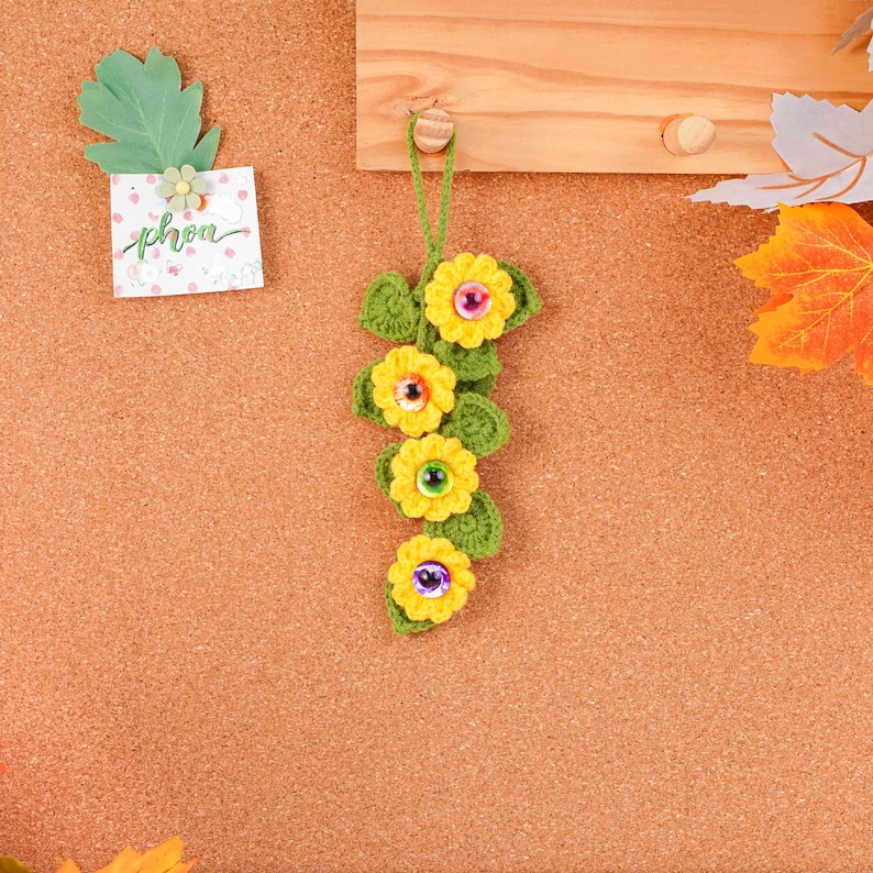 May include: A handmade crochet floral ornament featuring four yellow flowers with colorful centers and green leaves. The ornament is suspended by a green string and is attached to a wooden surface. A small card with the word "phon" is also visible.
