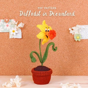 May include: A crocheted daffodil in a terracotta pot, with a yellow flower, green stem, and orange center. The text "PDF Pattern Daffodil in Dreamland" is above the flower. Decorative paper and flowers are pinned to the background.