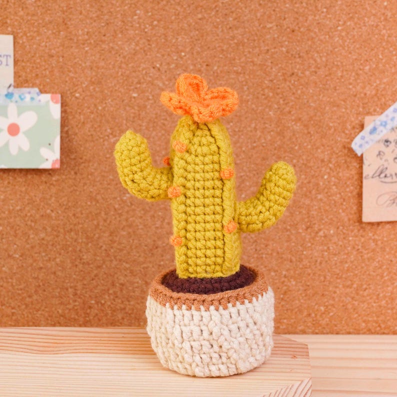May include: A crocheted cactus in a small pot. The cactus is yellow-green with small orange accents and a bright orange flower on top. It sits in a round, cream-colored crocheted pot with a brown rim, featuring a textured, hand-stitched appearance.