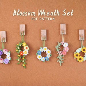 May include: Five handmade crochet blossom wreaths in different colours, suspended from wooden pegs on a brown surface. The text "Blossom Wreath Set PDF Pattern" is displayed above. Each wreath has a unique floral design.