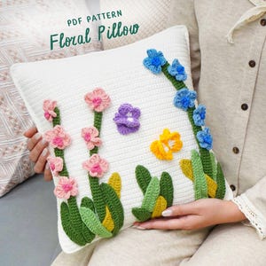 Könnte beinhalten: Ein weißes Häkelkissen mit bunten Blumenmustern. Das Kissen zeigt rosa, lila, gelbe und blaue Blumen mit grünen Stielen und Blättern. Der Text "PDF PATTERN Floral Pillow" ist oben zu sehen.