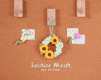 May include: A handmade crochet sunflower wreath with yellow and green accents, hanging against a corkboard background. The wreath is surrounded by small decorative cards and wooden hooks. The text "Sunshine Wreath" is displayed below.