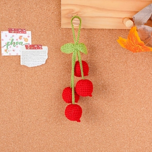 May include: A handmade crochet cherry decoration. The cherries are bright red, with a green stem and leaf. The decoration is hanging on a corkboard. The word "phoa" is visible on a small piece of paper.