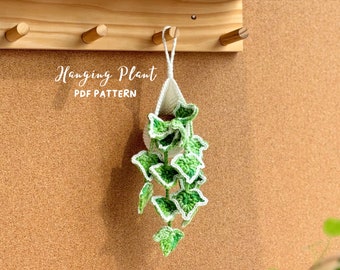 May include: A crocheted hanging plant with green leaves and white trim. The plant is hanging from a wooden rack with the text "Hanging Plant PDF Pattern" on a corkboard background.