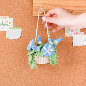 May include: A handmade crochet hanging basket with blue and white flowers and green leaves. The basket is suspended by a cream-colored string. The craft project is displayed against a corkboard background.