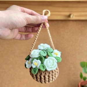 Op de afbeelding: Een gehaakte hangende plantenhanger met witte bloemen en groene bladeren. De plantenhanger is bruin en heeft een touwhandvat.