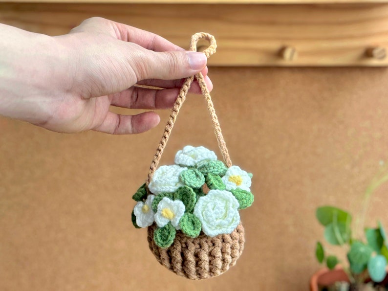 Op de afbeelding: Een gehaakte hangende plantenhanger met witte bloemen en groene bladeren. De plantenhanger is bruin en heeft een touwhandvat.
