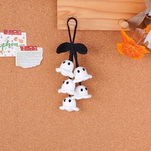 May include: A handmade Halloween decoration featuring five small, white crochet ghosts with black dot eyes. The ghosts are attached to a black crochet stem with a bow and a loop for hanging. The background is a corkboard.