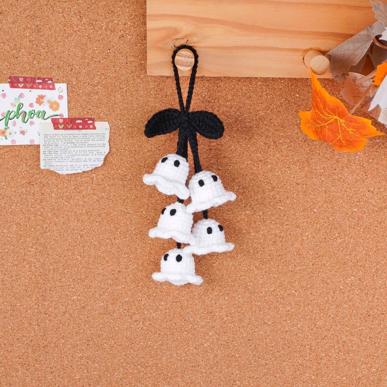 May include: A handmade Halloween decoration featuring five small, white crochet ghosts with black dot eyes. The ghosts are attached to a black crochet stem with a bow and a loop for hanging. The background is a corkboard.