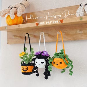 Puede incluir: Tres decoraciones colgantes de Halloween de ganchillo. Cada decoración es una maceta con forma de calabaza con un personaje diferente en su interior. Una tiene una calavera, otra una planta verde y la última flores moradas. Las decoraciones cuelgan de ganchos en una estantería de madera. La estantería tiene el texto "Halloween Hanging Decor PDF Pattern".