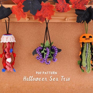 May include: Three crocheted hanging ornaments featuring a white eyeball jellyfish with red tentacles, a black pot with purple tentacles and green leaves, and an orange pumpkin jellyfish with green and black tentacles. Text reads: PDF PATTERN Halloween Sea Trio.