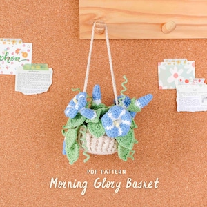 May include: A crocheted morning glory basket with blue and white flowers and green leaves hangs from a wooden board. The basket is beige and has a white cord for hanging. The text "PDF Pattern Morning Glory Basket" is below the basket. Decorative notes are pinned to the corkboard.