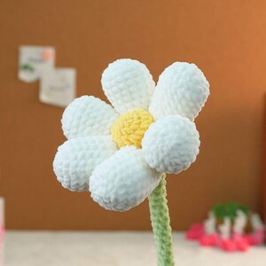 May include: A white crocheted daisy flower with a yellow center. The flower is attached to a green crocheted stem.