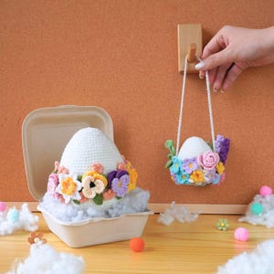 May include: Two crocheted Easter eggs decorated with colorful flowers. One egg is in a white paper container, and the other is hanging from a wooden hook.