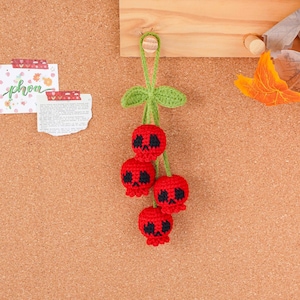May include: A handmade crochet charm featuring four red cherries with black skull faces. The cherries are attached to a green stem with leaves, hanging from a wooden peg. The charm is displayed against a corkboard background.