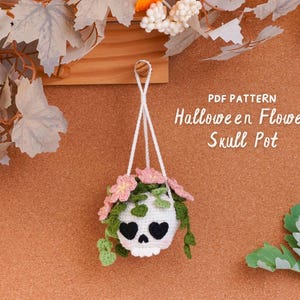 Könnte beinhalten: Ein weißer, gehäkelter, schädelförmiger Hängeübertopf mit schwarzen herzförmigen Augenhöhlen und rosa Blumen mit herabhängenden grünen Blättern. Der Text PDF PATTERN Halloween Flower Skull Pot ist rechts in Weiß aufgedruckt.