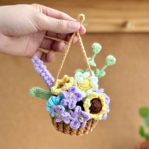 Op de afbeelding: Een geh&auml;kelde hangende bloemenmand met een verscheidenheid aan bloemen in tinten van paars, geel en wit. De mand is gemaakt van bruin garen en heeft een lus om op te hangen.