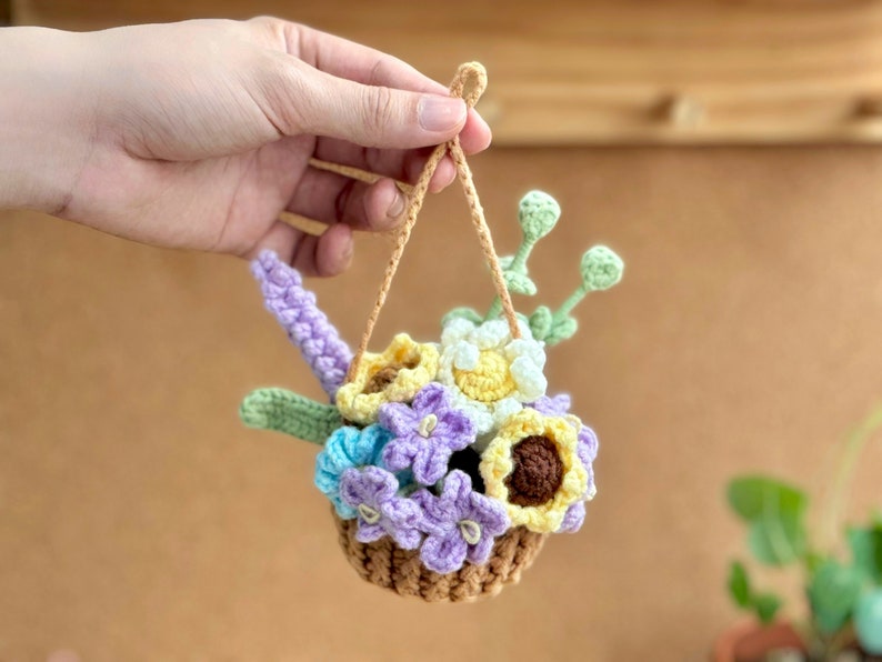 Op de afbeelding: Een geh&auml;kelde hangende bloemenmand met een verscheidenheid aan bloemen in tinten van paars, geel en wit. De mand is gemaakt van bruin garen en heeft een lus om op te hangen.