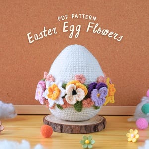 May include: A white crocheted Easter egg decorated with colorful crocheted flowers. The egg is sitting on a wooden base. The text "PDF PATTERN Easter Egg Flowers" is written above the egg.
