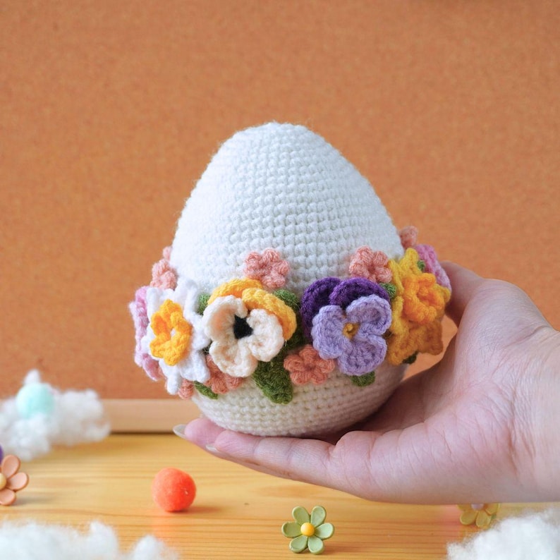 May include: A white crocheted egg decorated with a colorful flower garland. The garland features purple, yellow, pink, and white flowers.