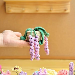 May include: A crocheted purple flower plant in a small brown pot with green leaves. The plant is held in a person's hand.