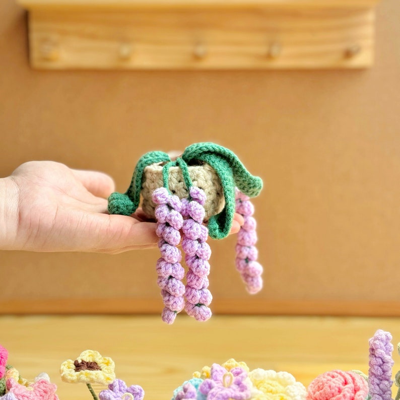 May include: A crocheted purple flower plant in a small brown pot with green leaves. The plant is held in a person's hand.