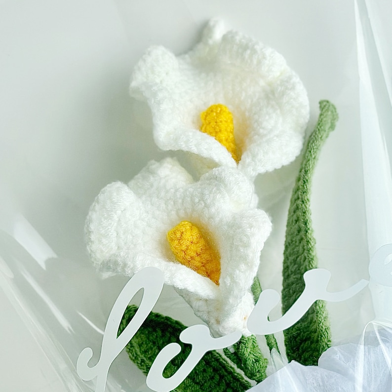 May include: A bouquet of two white crocheted calla lilies with yellow centers. The lilies are arranged with green crocheted leaves and the word "love" is written in white on the background.