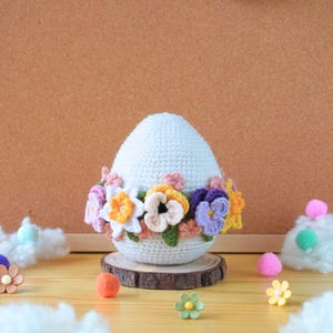 May include: A crocheted Easter egg with a white base and a colorful floral wreath around the middle. The egg is sitting on a wooden slice.