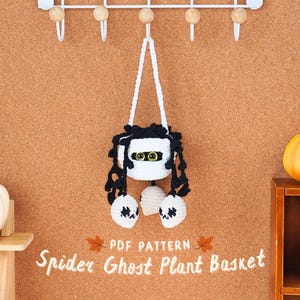 May include: A white crocheted ghost-shaped plant hanger with black spider legs and two white ghost-shaped ornaments hanging from the bottom. The hanger is hanging from a white hook on a corkboard wall. The text "PDF PATTERN Spider Ghost Plant Basket" is written below the hanger.