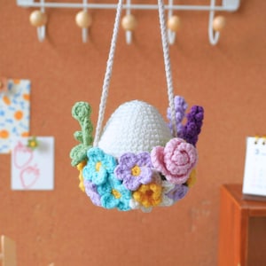 May include: A white crocheted egg with a floral wreath of pink, purple, blue, and yellow flowers hanging from a white string.