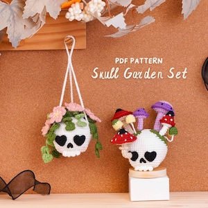 Könnte beinhalten: Eine Häkel-PDF-Anleitung mit dem Titel Skull Garden Set, die zwei weiße Totenköpfe zeigt. Einer ist ein hängender Übertopf mit rosa Blumen und grünen Ranken. Der andere steht auf einem weißen Block und ist mit roten, gelben und violetten Pilzen verziert.