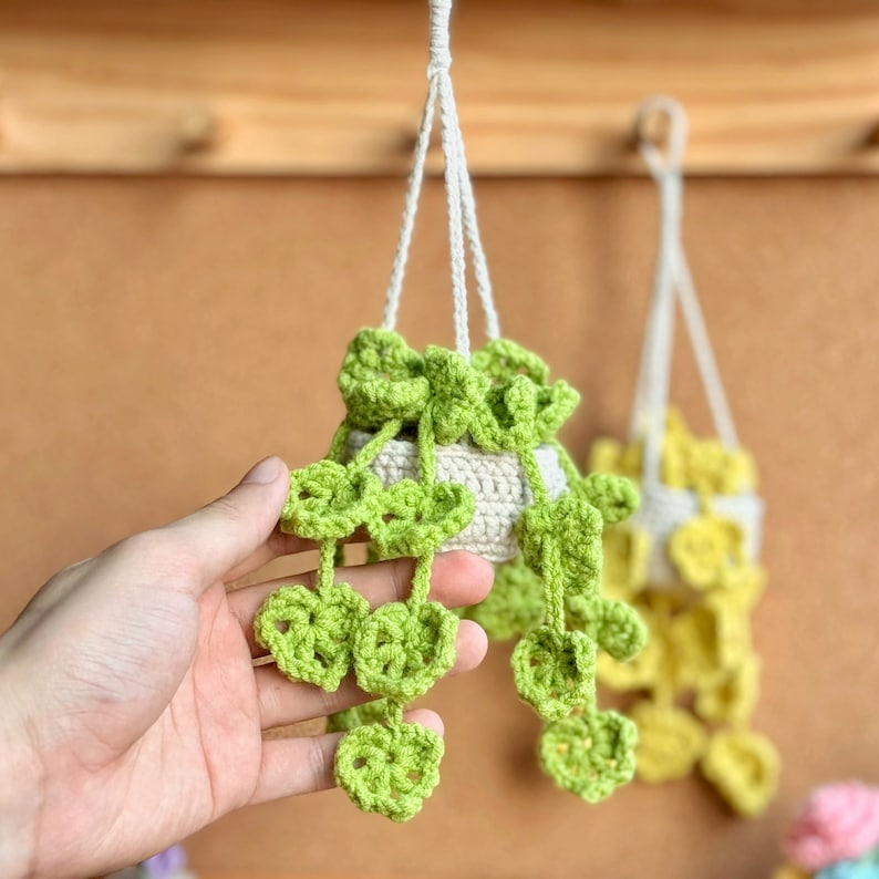 May include: Two crocheted hanging planters with green and yellow leaves. The planters are white and have a crocheted design. The planters are hanging from a wooden surface.