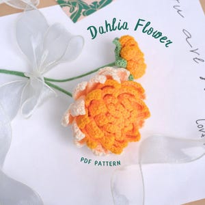 May include: A crocheted dahlia flower in shades of orange and white. The flower is on a white background with a sheer ribbon and the text "Dahlia Flower" written in black.