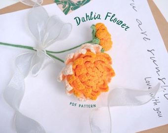 May include: A crocheted dahlia flower in shades of orange and white. The flower is on a white background with a sheer ribbon and the text "Dahlia Flower" written in black.