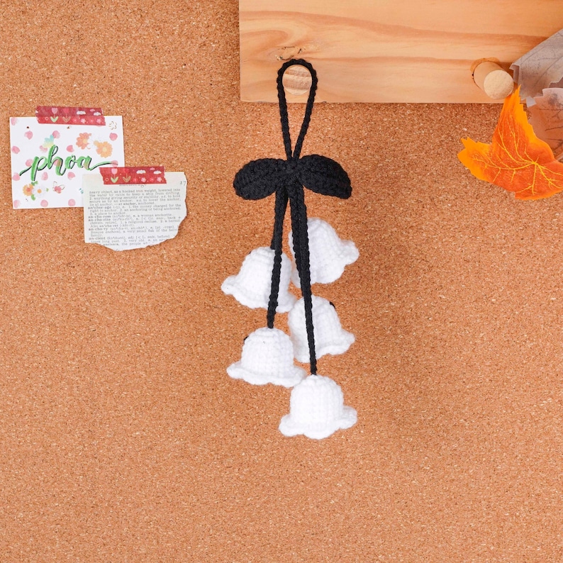 May include: A handmade crochet bell flower ornament. The ornament features five white bell-shaped flowers suspended from a black bow. The ornament is hanging on a corkboard. The word "phoa" is written on a small card.