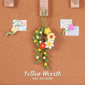 Puede incluir: Una corona amarilla de ganchillo hecha a mano con flores blancas, naranjas y amarillas, hojas verdes y pequeñas bolas blancas. La corona está colgada en un corcho. El texto "Yellow Wreath PDF Pattern" está en la parte inferior.