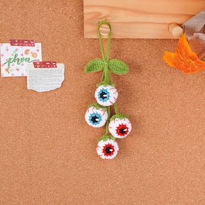 May include: A handmade crochet Halloween decoration featuring four eyeball ornaments. Each eyeball is white with red veins, a black pupil, and either blue or red iris. The eyeballs are connected by green stems and leaves.