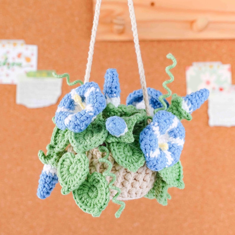 May include: A handmade crochet hanging basket with blue and white morning glory flowers, green leaves, and a beige pot. The basket is suspended by a white cord. The flowers have yellow centers. The overall theme is home decor.