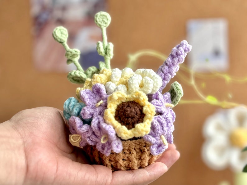 Op de afbeelding: Een gehaakt bloemstuk in een bruine mand. Het arrangement bevat paarse, gele en witte bloemen, evenals groene bladeren.