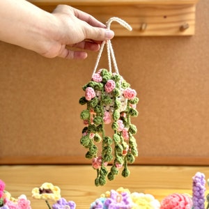May include: A crocheted hanging planter with green leaves and pink flowers. The planter is made of yarn and is hanging from a string.