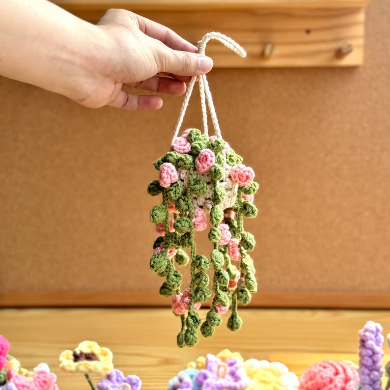 May include: A crocheted hanging planter with green leaves and pink flowers. The planter is made of yarn and is hanging from a string.
